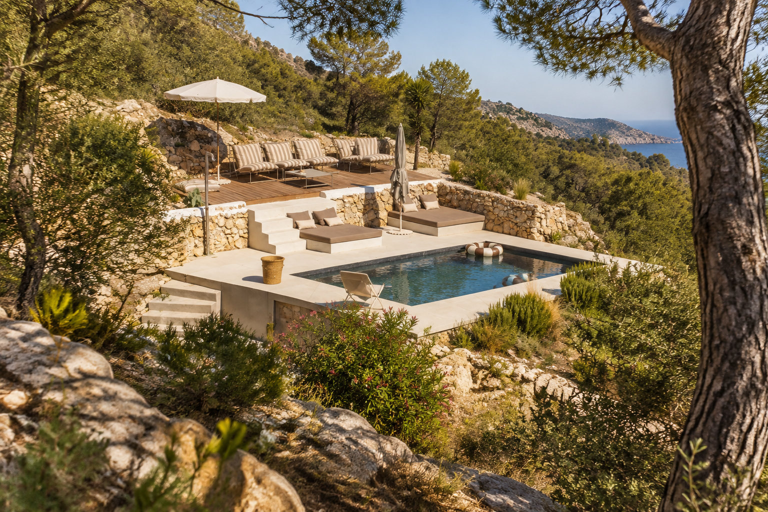 Une piscine entourée de terrasses en pierre, de banquettes et de parasols se trouve au milieu d’un paysage méditerranéen arboré, avec la mer à l’arrière-plan.